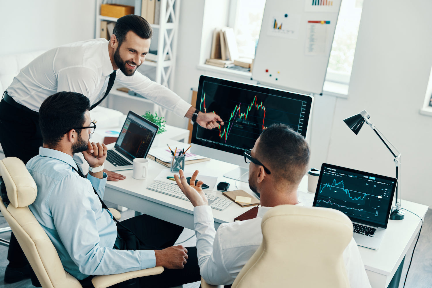 Three professionals in a modern office setting analyzing financial data on computer screens.